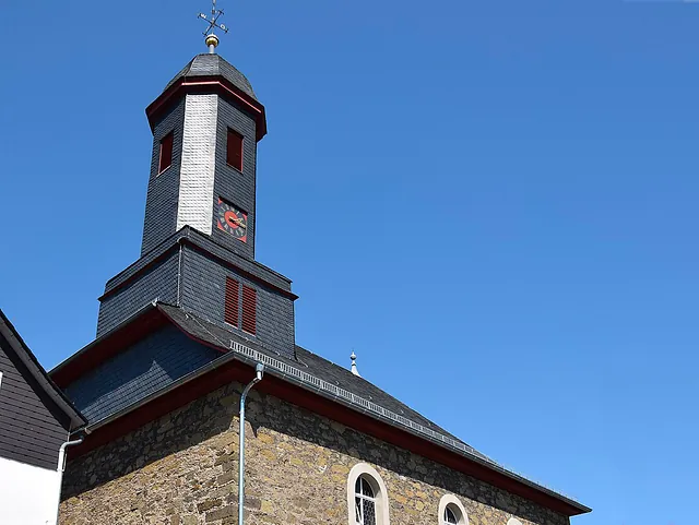 Nahaufnahme eines dunklen Kirchturms mit rotem Zifferblatt vor blauem Himmel.