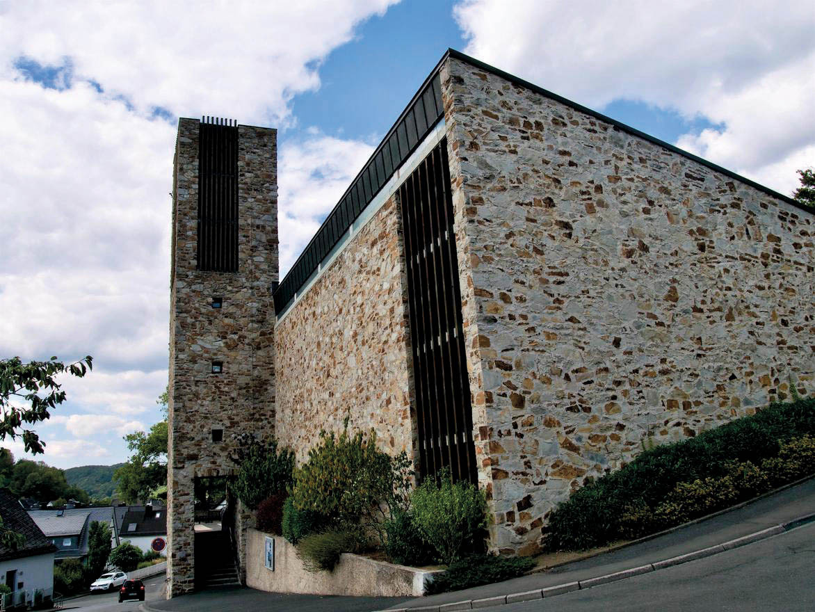 Moderne Kirche aus Bruchstein mit hohem, schmalem Glockenturm an einem Hang.