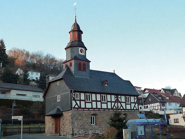 Kirche mit Fachwerkobergeschoss und Turm in einer dörflichen Umgebung.