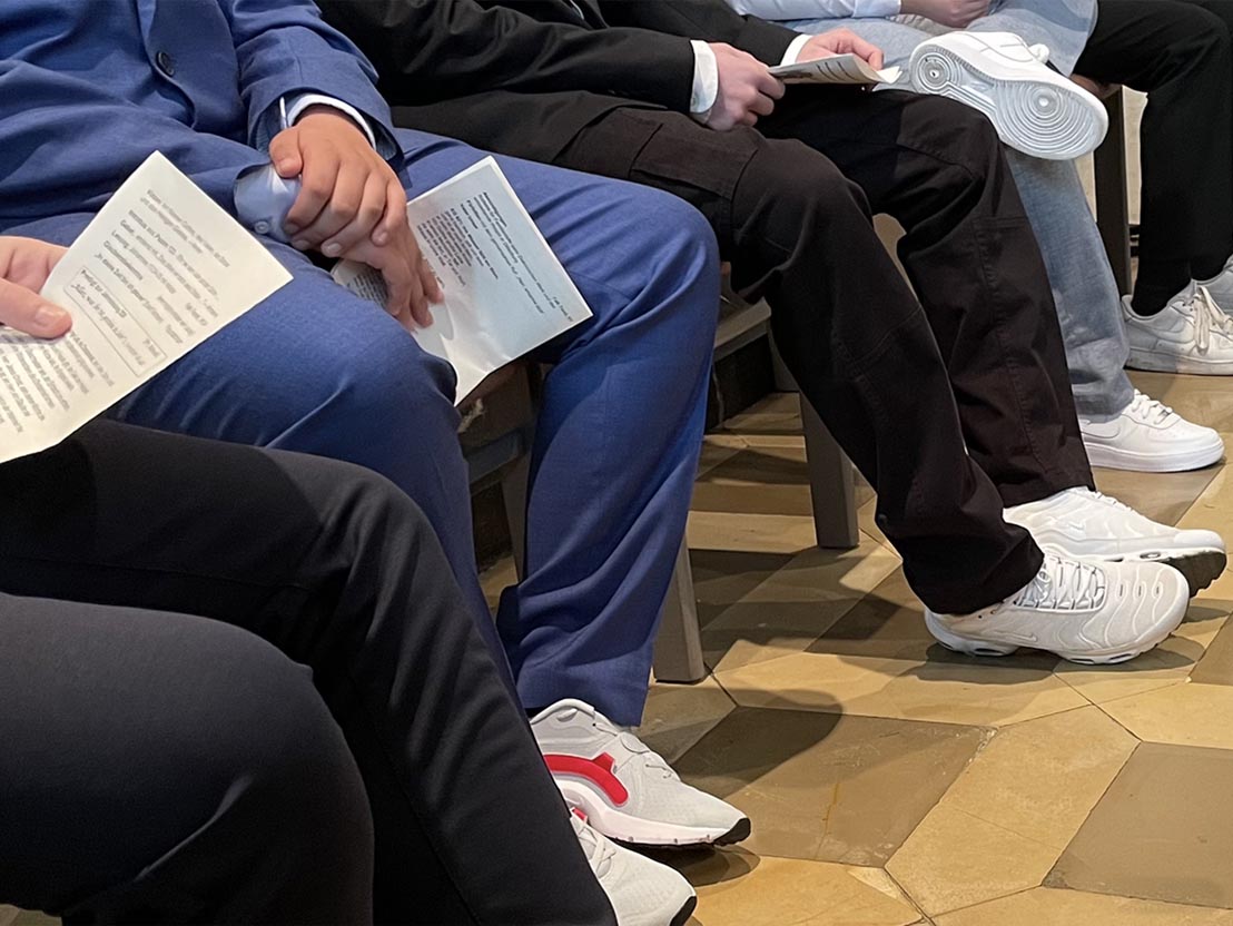 Sitzende Personen in festlicher Kleidung halten Programme in den Händen.